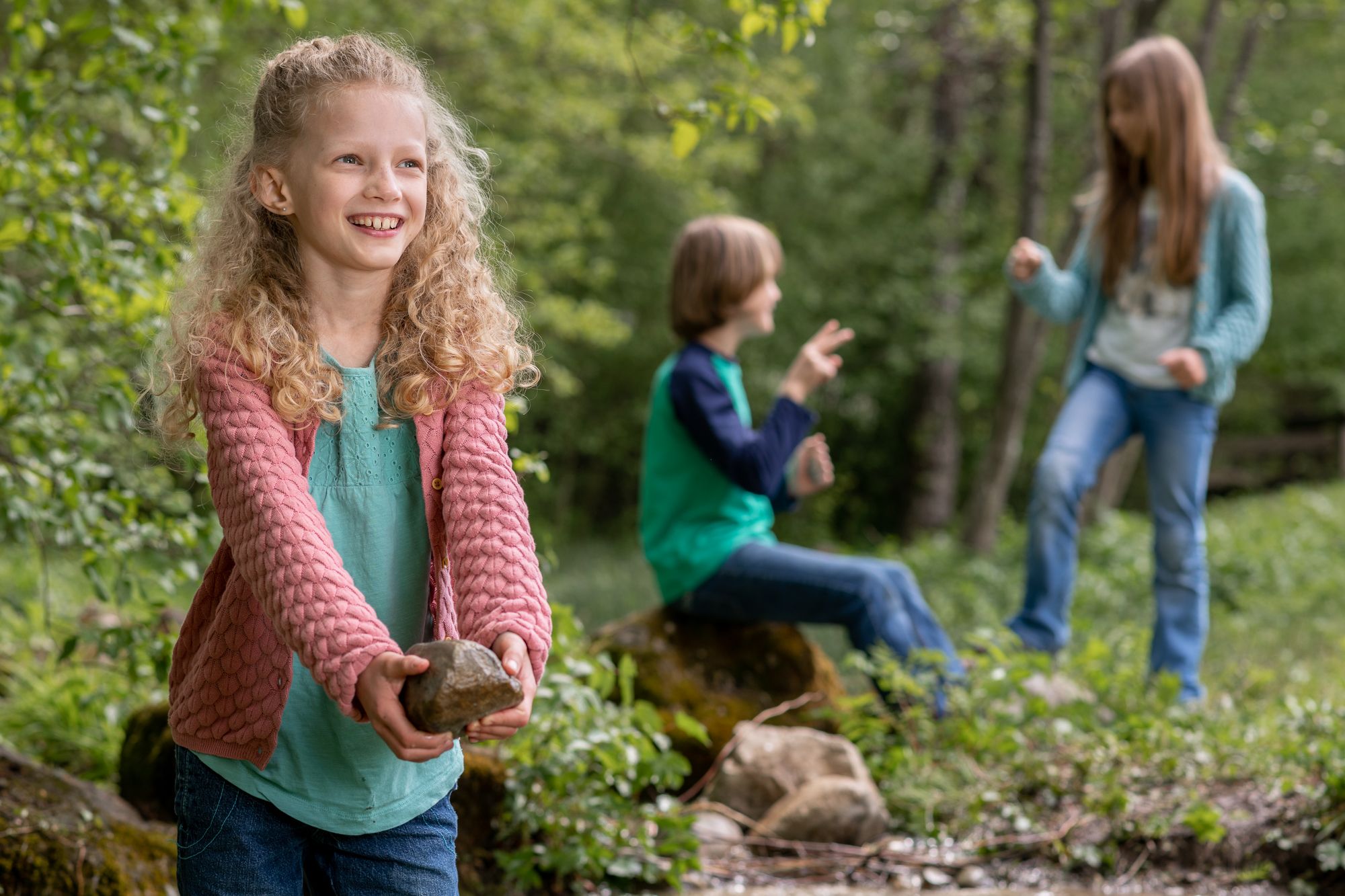 A smiling girl in nature holding a rock and two children playing in the background